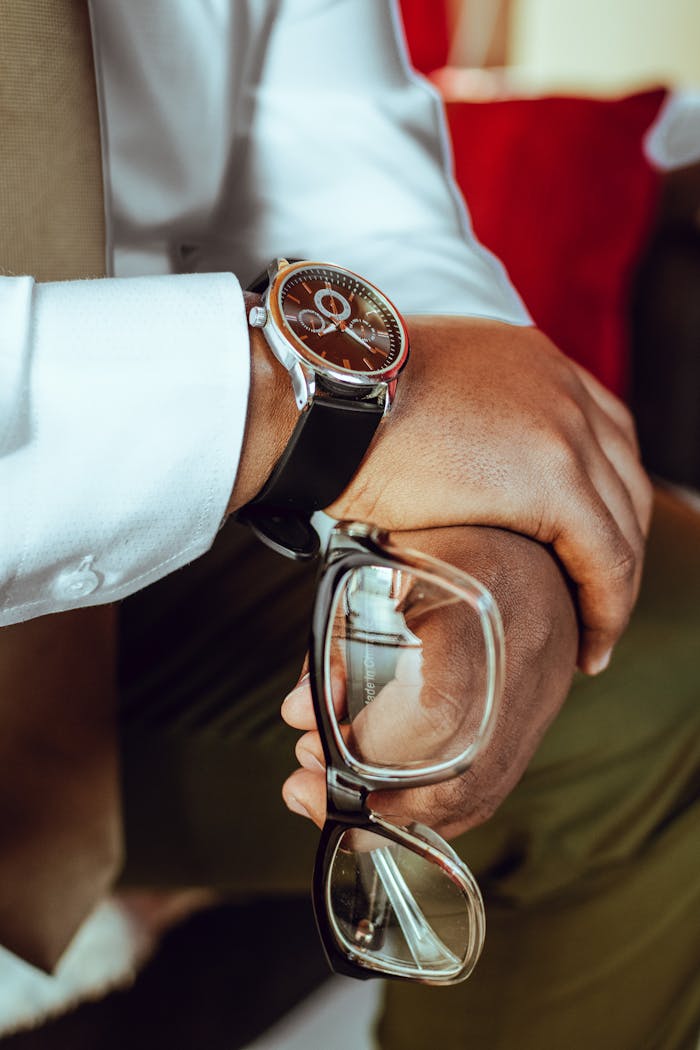 Close-up shot of a man in formal attire holding eyeglasses, showcasing a stylish wristwatch.